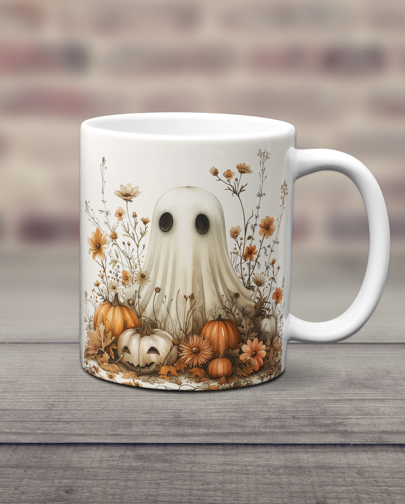 K&ouml;nnte beinhalten: Wei&szlig;e Keramiktasse mit einer Geisterillustration, umgeben von orangefarbenen K&uuml;rbissen und Herbstlaub. Der Geist hat zwei schwarze Augen und einen wei&szlig;en, lakenartigen K&ouml;rper.