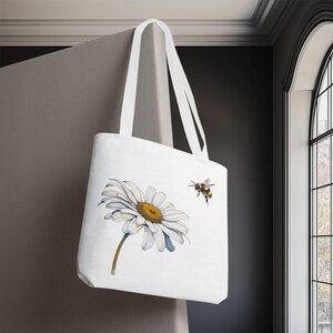 Op de afbeelding: Witte draagtas met een grote aquarel madelief met een gele kern en witte bloemblaadjes, en een hommel. De tas heeft witte handvatten en hangt tegen een grijze achtergrond.