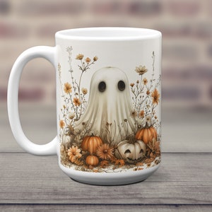 K&ouml;nnte beinhalten: Wei&szlig;e Keramiktasse mit einer Geisterillustration, umgeben von orangefarbenen K&uuml;rbissen und Herbstblumen. Der Geist hat zwei schwarze Augen und einen wei&szlig;en, lakenartigen K&ouml;rper.