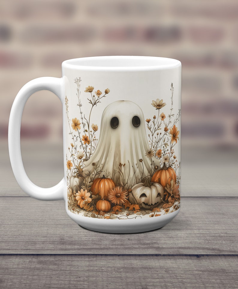 K&ouml;nnte beinhalten: Wei&szlig;e Keramiktasse mit einer Geisterillustration, umgeben von orangefarbenen K&uuml;rbissen und Herbstblumen. Der Geist hat zwei schwarze Augen und einen wei&szlig;en, lakenartigen K&ouml;rper.