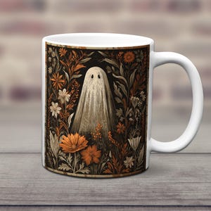 May include: White ceramic mug with a vintage illustration of a ghost amidst orange, brown, and white flowers on a black backdrop. The mug has a white handle. The mug is set on a wooden surface.