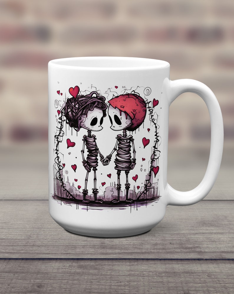 May include: White ceramic coffee mug with a graphic of two skeletons holding hands. The skeletons are wrapped in bandages and have hearts around them. The mug has a black handle.