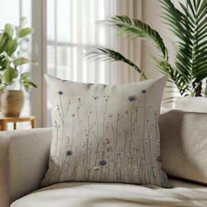 May include: A decorative throw pillow with a floral design. The pillow features a cream-colored background with a pattern of delicate wildflowers in shades of white, blue, and brown. The pillow is placed on a beige sofa.