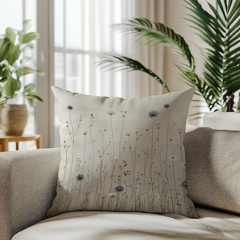 May include: A decorative throw pillow with a floral design. The pillow features a cream-colored background with a pattern of delicate wildflowers in shades of white, blue, and brown. The pillow is placed on a beige sofa.