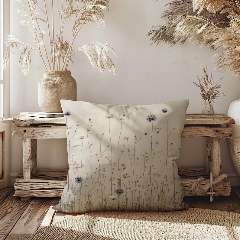 May include: A square decorative pillow with a floral design. The pillow features a cream-colored background with delicate illustrations of wildflowers in shades of blue, white, and yellow. The pillow is placed on a rustic wooden bench, adding a touch of nature-inspired decor.