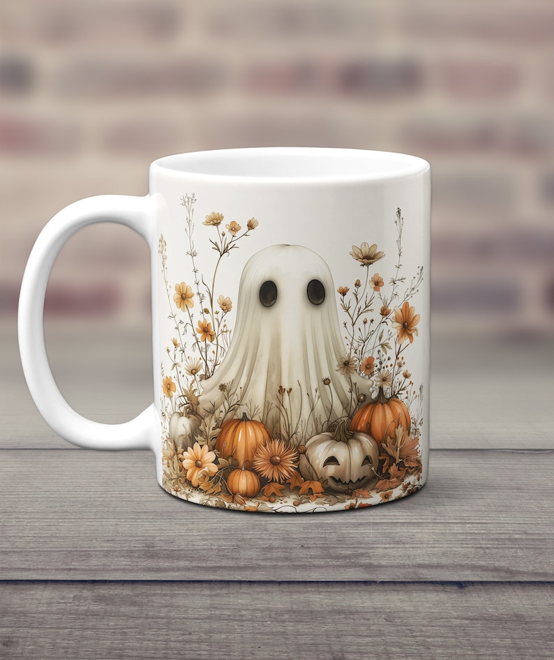 K&ouml;nnte beinhalten: Wei&szlig;e Keramiktasse mit einer Geisterillustration, umgeben von orangefarbenen und gelben Blumen und K&uuml;rbissen. Der Geist hat schwarze Augen und einen wei&szlig;en, lakenartigen K&ouml;rper.