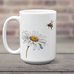 May include: White ceramic mug with a white daisy and a bumblebee in flight. The daisy has a yellow center and white petals.