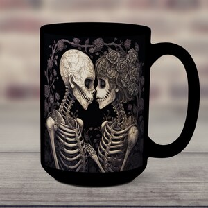 May include: Black coffee mug with a graphic of two skeletons embracing, surrounded by roses and vines. The skeletons are wearing floral crowns and are looking at each other.