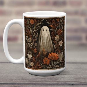May include: White ceramic mug with a vintage illustration of a ghost in a floral garden. The design features orange and white flowers on a black background. The mug has a large handle and is set against a wooden surface.