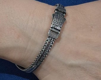 Puede incluir: Una pulsera de plata con un diseño intrincado. La pulsera presenta una cadena tejida y un cierre decorativo con patrones detallados. El metal tiene un aspecto ligeramente oxidado, lo que le da un aspecto vintage. La pulsera se lleva en la muñeca.