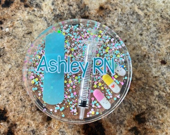 May include: A round, clear resin coaster with a light blue bandage, a syringe, and colorful pills. The coaster has a glittery, confetti-like design and the text "Ashley RN" is written on it.