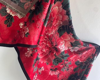 May include: A red scarf with a floral design, showcasing large flowers in red, pink, and white, on a dark green and black backdrop. The scarf has a black border and a fringed edge.