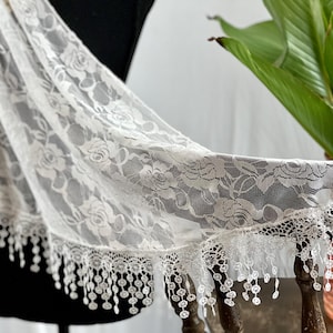May include: White lace fabric with a floral pattern and a fringed edge. The fabric is draped over a mannequin and a plant is visible in the background.
