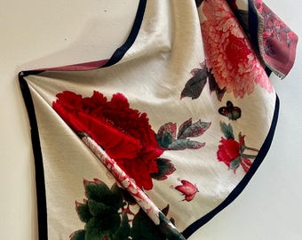 May include: A cream scarf with a large red floral print and dark navy blue trim. The scarf features a repeating pattern of red roses and green leaves. The edges are finished with a fringe.