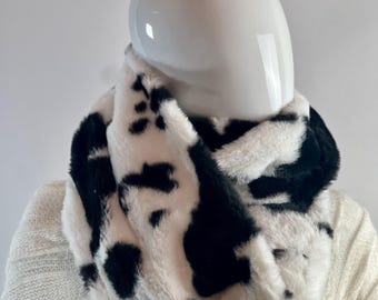May include: A white and black faux fur infinity scarf. The scarf is made of soft, fluffy material and has a cow print pattern.