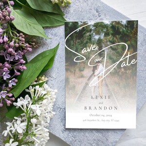 May include: Save the date card with a blurred photo of a couple in a forest setting. The text reads "Save our Date" with a white script font. The couple's names are Lexie and Brandon, with the date October 04, 2024, and the address 438 Anywhere St., Avy city, ST 0145.