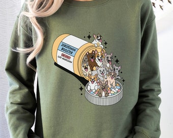 May include: Military green sweatshirt featuring a cartoon pill bottle labeled "Serotonin Booster" with the phrase "Cute Aggression." The bottle is overflowing with cartoon dogs of various breeds. The sweatshirt is a solid color.