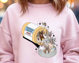 May include: A pink sweatshirt with a graphic of a pill bottle labeled "Serotonin Booster" with a bunch of cartoon dogs spilling out of it.