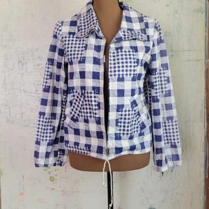 May include: A long-sleeved zip-up jacket featuring a blue and white gingham check pattern with a patchwork design. The jacket includes a collar, a full-length front zipper, and a drawstring hem with white cords at the bottom for an adjustable fit.