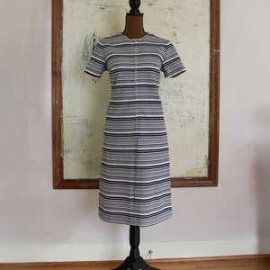 Puede incluir: Un vestido a rayas blancas y azul marino con una cremallera en la parte delantera. El vestido está en un maniquí negro contra una pared blanca.