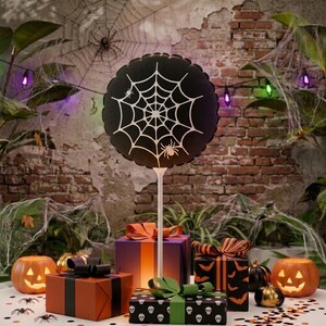 Könnte beinhalten: Ein runder schwarzer Ballon mit weißem Spinnennetz- und Spinnenmotiv, befestigt an einem weißen Stab. Der Ballon ist zwischen mehreren Geschenkboxen in Orange, Lila und Schwarz sowie zwei orangefarbenen Kürbislaternen platziert.