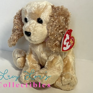 May include: A beige and brown plush toy dog with a pink bow. The dog is sitting with its head tilted to the side. The dog has a red and white Ty tag attached to its ear that says "Original Beanie Babies".