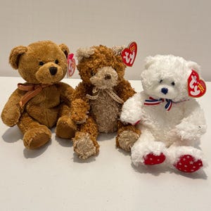 May include: Three plush teddy bears, one brown, one light brown, and one white. The brown bear has a brown ribbon around its neck. The light brown bear has a tan ribbon around its neck. The white bear has a red, white, and blue ribbon around its neck. All three bears have Ty tags attached.