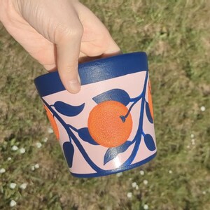 May include: A hand-painted ceramic flower pot with a navy blue rim and a light pink body. The pot is decorated with orange citrus fruit and navy blue leaves and branches.