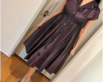 May include: A vintage-style, knee-length dress in a shimmering, dark purple fabric. The dress features a collared neckline, short sleeves, and a button-down front. The skirt is flared, and the dress is paired with silver pointed-toe shoes.