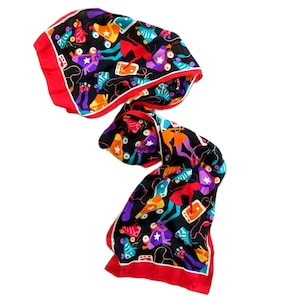 Puede incluir: Una bufanda negra con un vibrante diseño de patines en rojo, naranja, morado y turquesa. La bufanda tiene un borde rojo y un ribete blanco. El diseño incluye estrellas, cintas de cassette y figuras en movimiento.