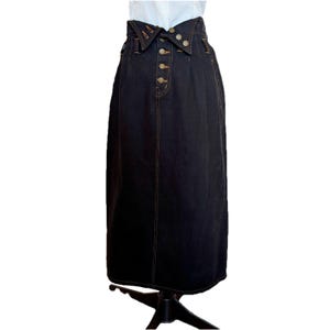 May include: A dark wash denim skirt with a high waist and button-down front. The skirt features a unique collar-like waistband and decorative stitching. The skirt is a midi length, and the stitching is a contrasting gold color.