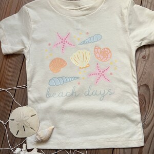 K&ouml;nnte beinhalten: Ein cremefarbenes T-Shirt mit einem Aufdruck von Muscheln, Seesternen und dem Text "beach days" in hellblau.