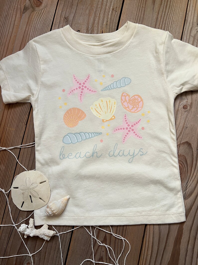 K&ouml;nnte beinhalten: Ein cremefarbenes T-Shirt mit einem Aufdruck von Muscheln, Seesternen und dem Text "beach days" in hellblau.