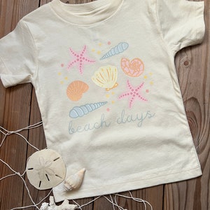 K&ouml;nnte beinhalten: Ein cremefarbenes T-Shirt mit einem Aufdruck von Muscheln, Seesternen und dem Text "beach days".