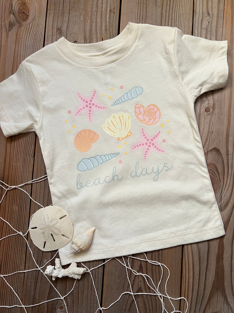 K&ouml;nnte beinhalten: Ein cremefarbenes T-Shirt mit einem Aufdruck von Muscheln, Seesternen und dem Text "beach days".