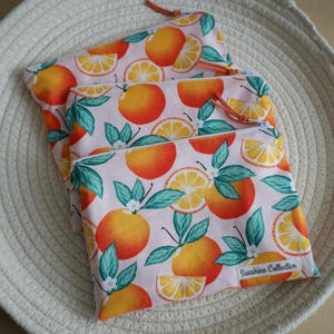 May include: Three pink zippered pouches with a white background and a repeating pattern of orange slices and green leaves. The pouches have a white label with the text "Sunshine Collective."