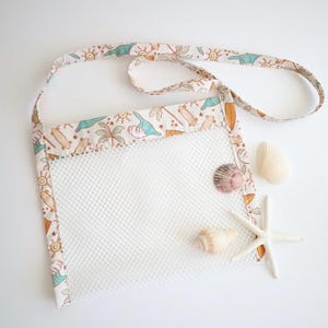 May include: A white mesh beach bag with a patterned strap featuring surfboards, palm trees, and dinosaurs. The bag is decorated with seashells and a starfish, perfect for collecting treasures at the beach.