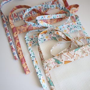 May include: Several reusable mesh produce bags with colorful fabric trim featuring seashell, starfish, and lobster patterns. The bags have long straps for carrying and are arranged on a white surface. The bags are designed for carrying groceries.