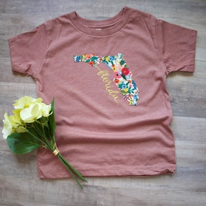 May include: A mauve t-shirt with a floral print of the state of Florida and the word "Florida" in gold script.