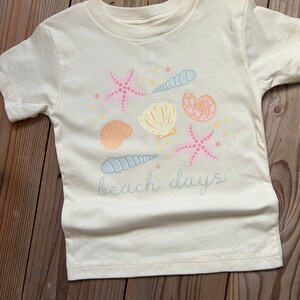 K&ouml;nnte beinhalten: Ein wei&szlig;es T-Shirt mit einem Strandmotiv mit rosa und blauen Muscheln, Seesternen und dem Text "beach days".