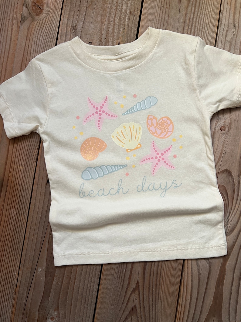 K&ouml;nnte beinhalten: Ein wei&szlig;es T-Shirt mit einem Strandmotiv mit rosa und blauen Muscheln, Seesternen und dem Text "beach days".