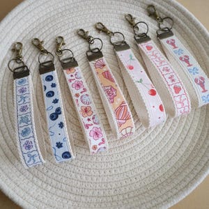 May include: A collection of fabric keychains with various patterns, including flowers, cherries, and lobsters. Each keychain has a metal clasp and a patterned fabric strap with a white border. The keychains are arranged on a woven, circular tray.