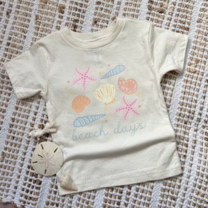 K&ouml;nnte beinhalten: Ein cremefarbenes T-Shirt mit kurzen &Auml;rmeln und einem Aufdruck von Muscheln und Seesternen. Der Text "beach days" ist auf das Shirt gedruckt.
