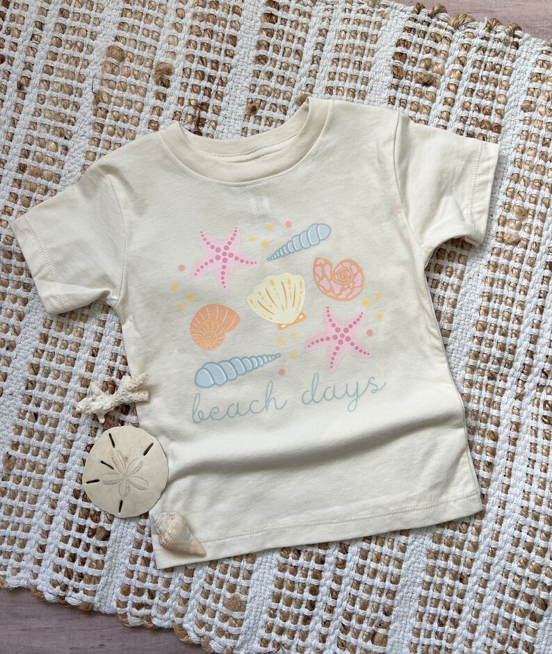 K&ouml;nnte beinhalten: Ein cremefarbenes T-Shirt mit kurzen &Auml;rmeln und einem Aufdruck von Muscheln und Seesternen. Der Text "beach days" ist auf das Shirt gedruckt.