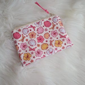 May include: A small, rectangular zippered pouch with a white background and a repeating pattern of pink and yellow seashells, flowers, and bows. The zipper pull is pink, and the pouch is likely made of fabric.