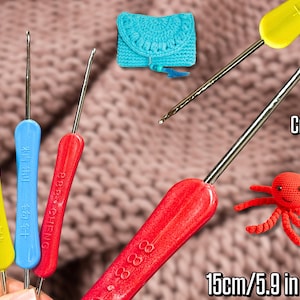 Pode incluir: Agulhas de crochê em amarelo, azul e vermelho com pontas de metal prateado. A imagem inclui um pequeno polvo de crochê e uma bolsa de crochê turquesa. O texto "Crochet Hook" e "15cm/5.9 in" também são visíveis.