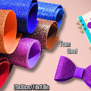 Puede incluir: Hojas de espuma con purpurina enrolladas en rojo, naranja, azul, marrón y morado. También se ve un lazo morado y un cuaderno con una decoración floral. Se muestra el texto "Foam Sheet" y "20x30cm/7.8x11.8in".