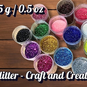 Op de afbeelding: Een verzameling glitter in verschillende kleuren, waaronder roze, groen, blauw en goud, weergegeven in kleine, transparante containers. De afbeelding bevat de tekst "15 g / 0.5 oz" en "Glitter - Craft and Create". De glitter ligt op een houten oppervlak.