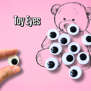 Puede incluir: Una colección de ojos de juguete con pupilas negras y contornos blancos, algunos colocados sobre un boceto de oso de peluche rosa. Un ojo es sostenido por una mano. Las palabras "Toy Eyes" están escritas en blanco y negro.