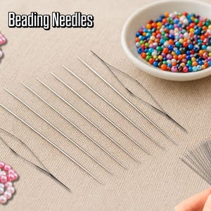 Op de afbeelding: Een assortiment rijgnaalden, een kleine kom met kleurrijke kralen en twee strengen roze en witte kralen worden gepresenteerd op een beige oppervlak. De tekst "Beading Needles" staat bovenaan. Een spoel garen is ook zichtbaar.